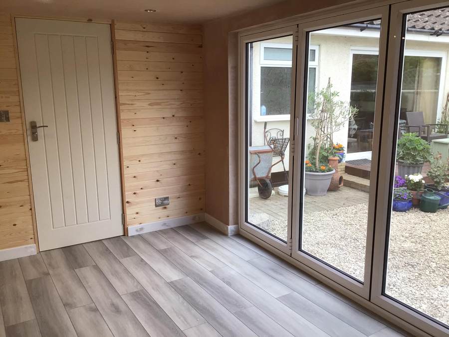 Converted garage with wood-effect flooring, timber panelled wall and sliding patio doors opening onto the garden